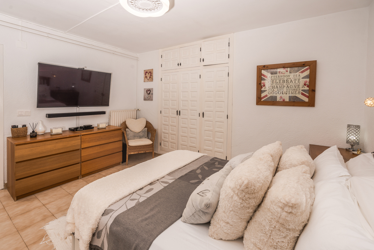 Slaapkamer met witte inbouwkasten, een grote wand-tv boven een houten dressoir en tegelvloer. Voorzien van een radiator en een rieten fauteuil.