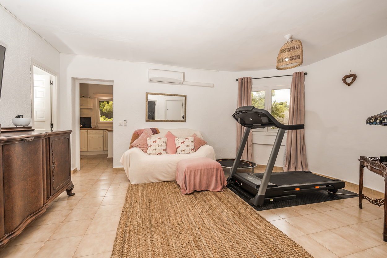 Veelzijdige woonkamer met fitnesshoek, loopband, tegelvloer en airconditioning, met een open verbinding naar de aangrenzende keuken.