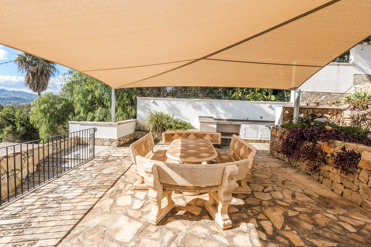 Buiten eethoek met een bewerkte stenen tafel en banken, overschaduwd door een beige schaduwdoek op een terras van natuursteen naast de buitenkeuken.