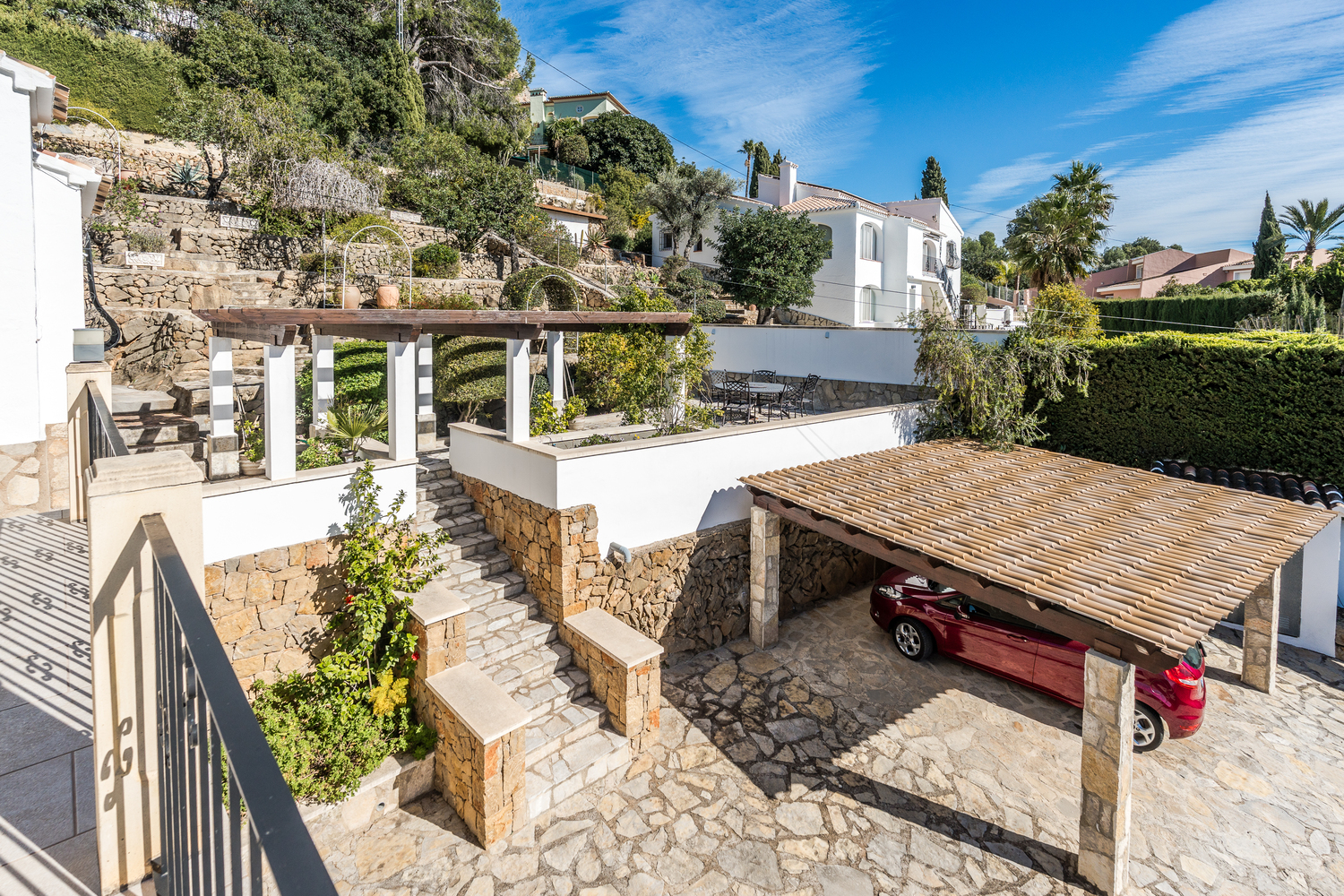 Buitenaanzicht met een bestrate oprit, een overdekte carport met pannendak en een terrasvormige mediterrane tuin met natuurstenen muren.