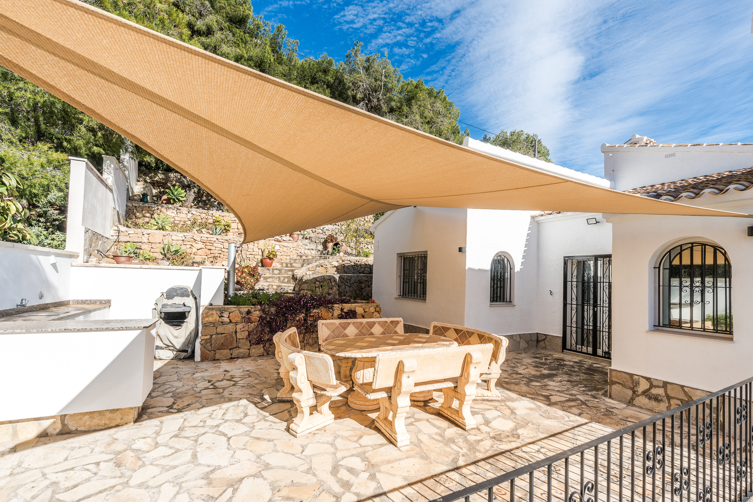 Buitenterras met een stenen eethoek onder een groot schaduwdoek, omgeven door natuurstenen muren en mediterrane beplanting bij de witgestuukte villa.