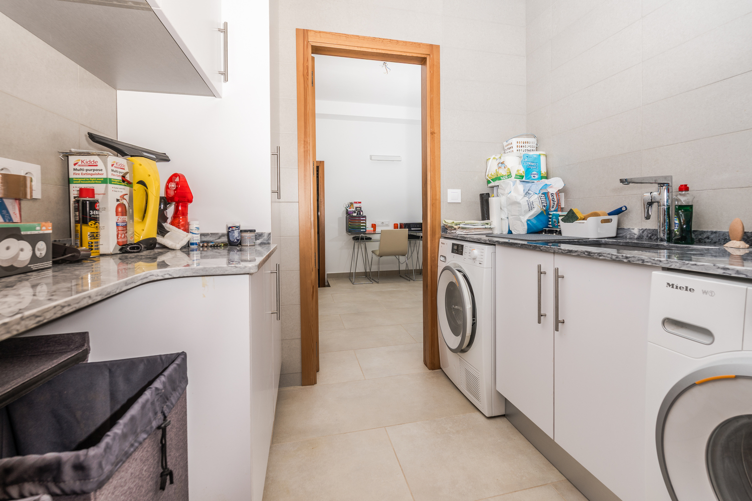 Functionele wasruimte met witte kasten, grijs marmeren werkbladen en Miele-apparatuur, met uitzicht op de aangrenzende kantoorruimte.