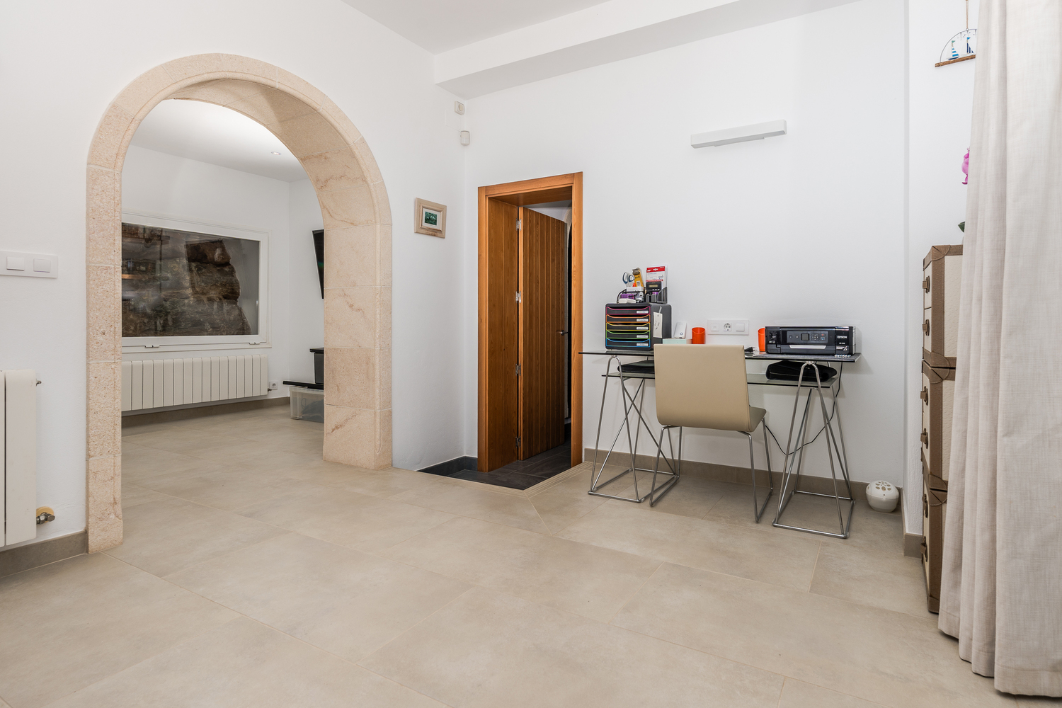 Kantoorruimte met een glazen bureau en een traditionele Tosca stenen boog die toegang biedt tot een kamer met neutrale tegelvloer.