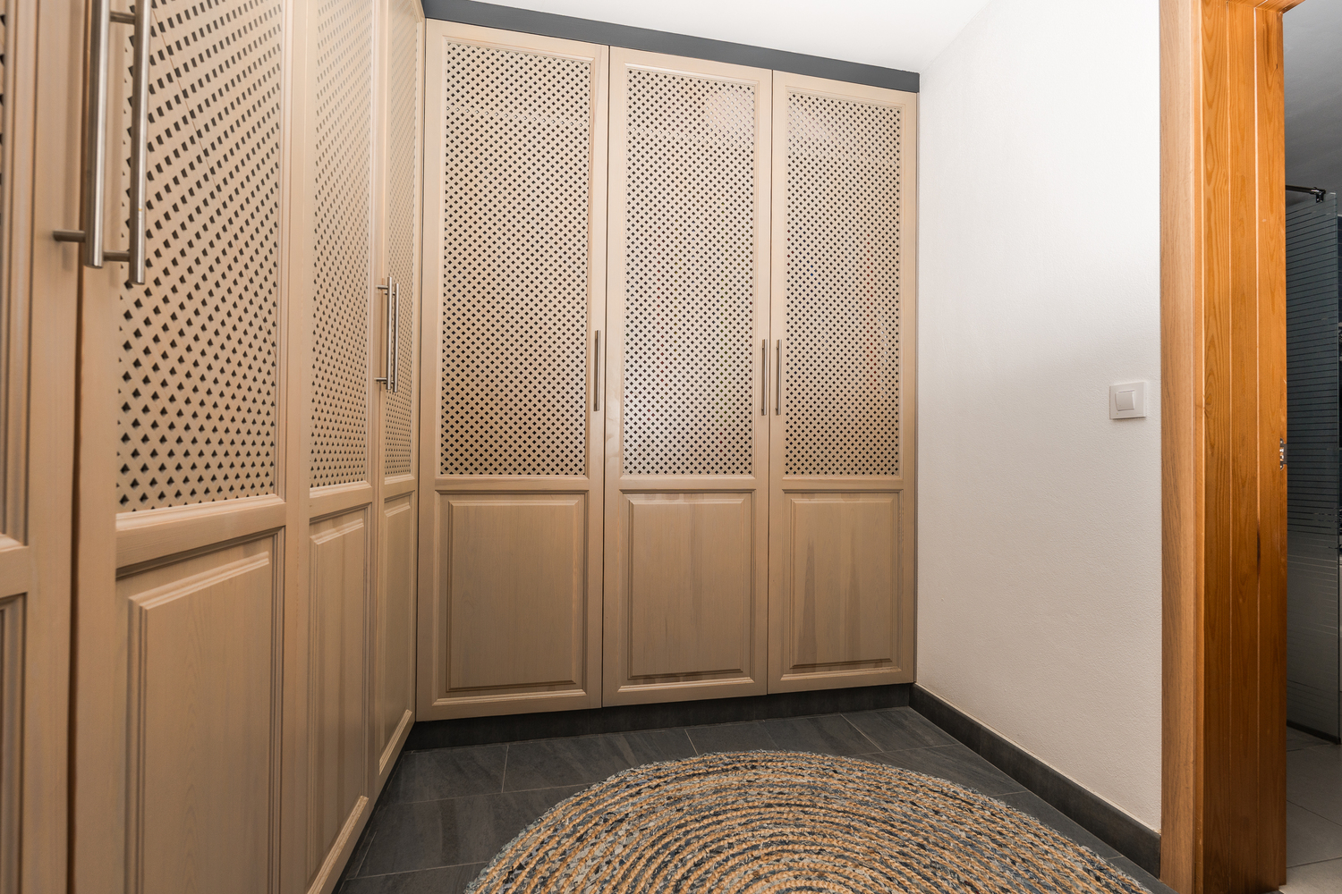 Kleedkamer met ingebouwde houten kasten voorzien van decoratieve roosterpanelen, donkere tegelvloer en een rond geweven vloerkleed.