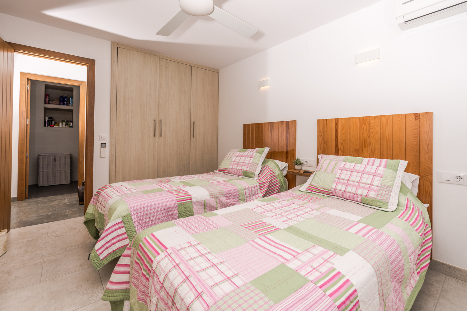 Lichte slaapkamer met twee eenpersoonsbedden, houten hoofdborden, inbouwkasten, plafondventilator en toegang tot een aangrenzende badkamer.