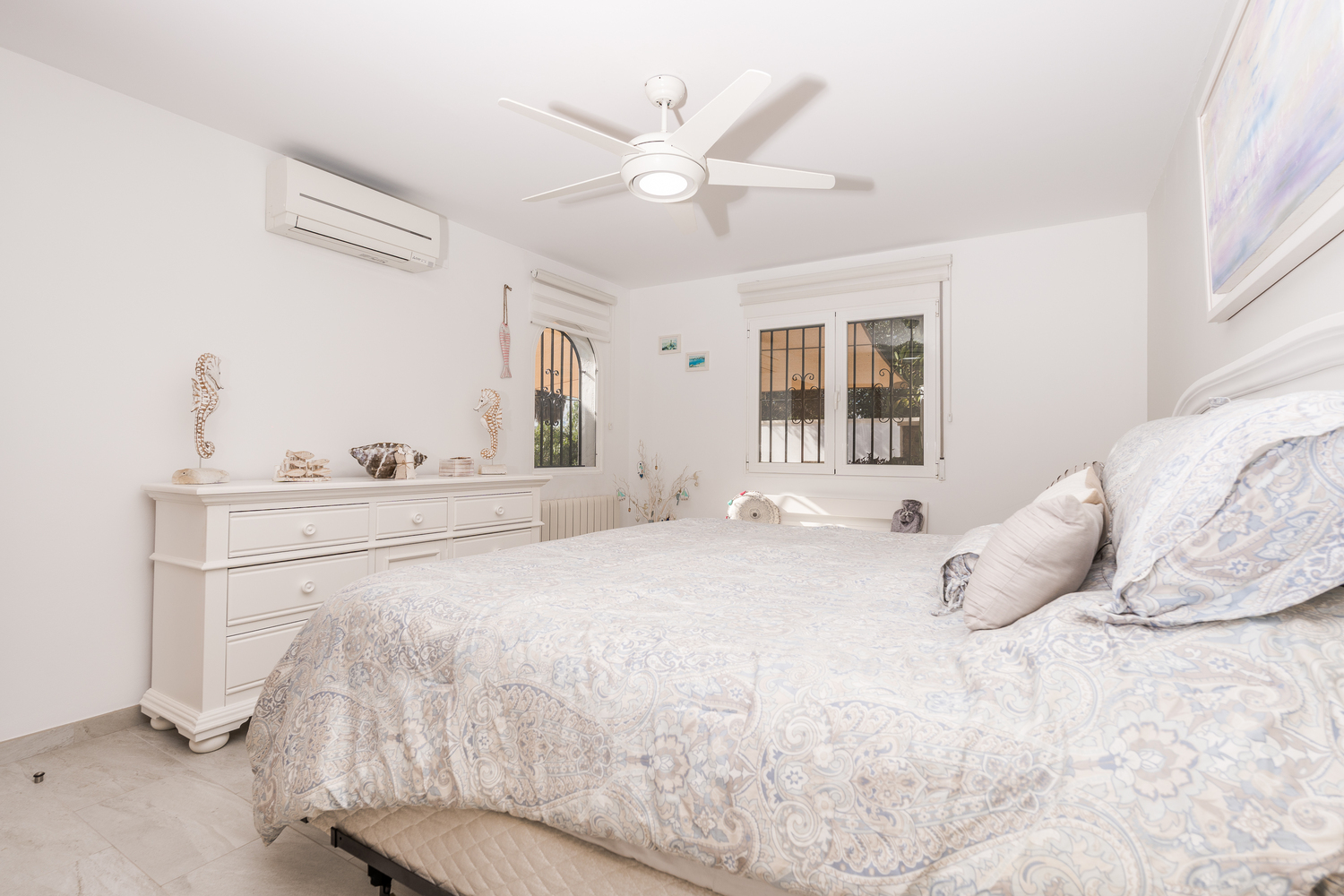 Lichte slaapkamer met witte muren, een plafondventilator, airconditioning en een witte ladekast. Voorzien van ramen met veiligheidstralies en een tegelvloer.