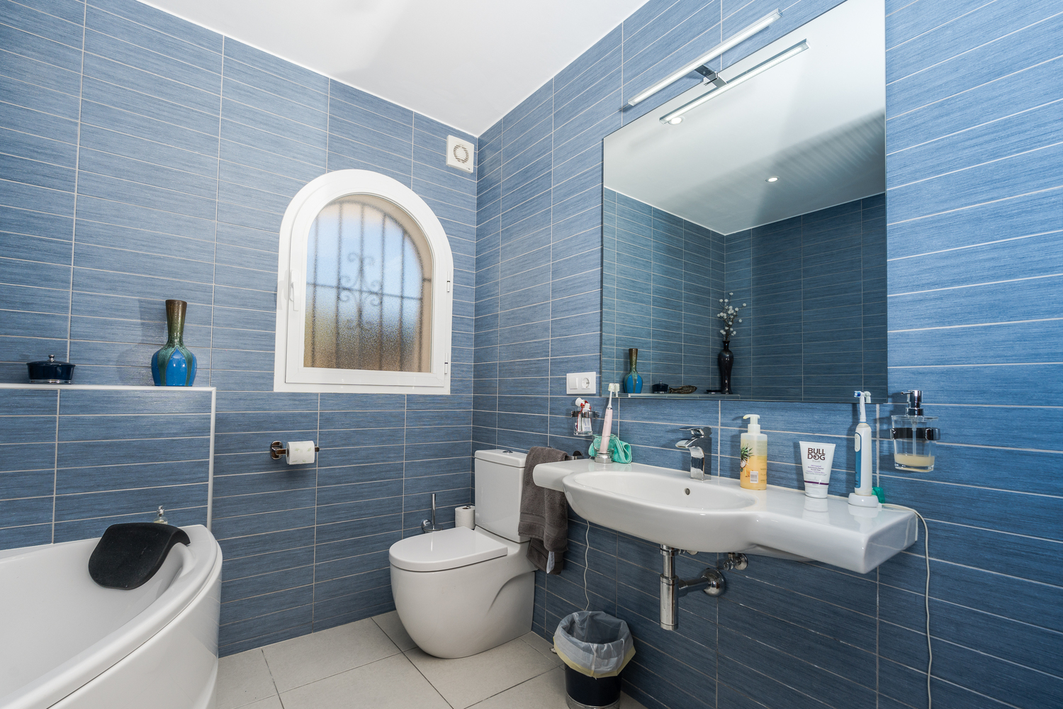 Moderne badkamer met blauwe textuurtegels, een boogvenster met matglas, wit keramisch wastafelmeubel en een grote spiegel met bovenverlichting.