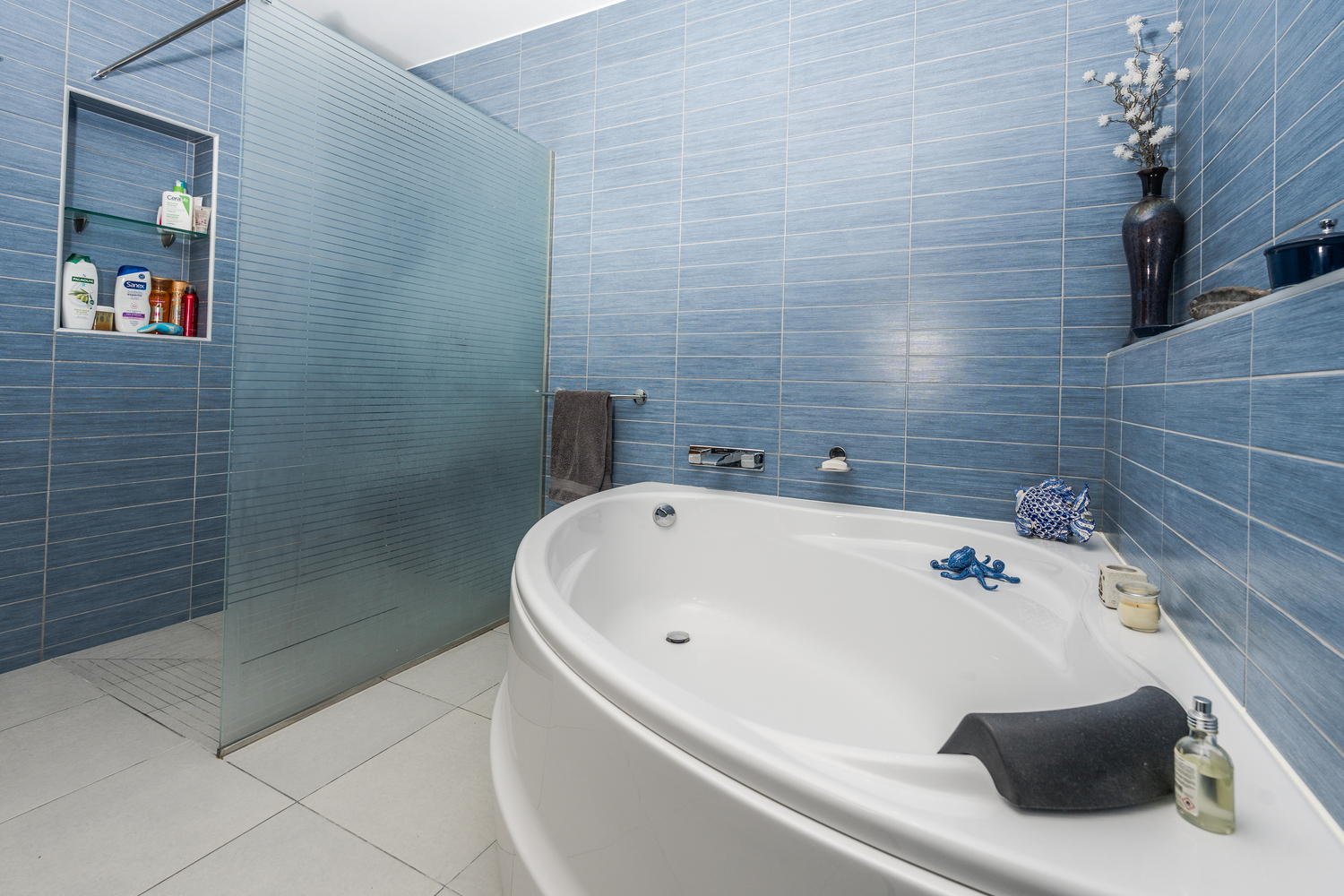 Moderne badkamer met een groot ovaal ligbad en een inloopdouche met matglazen wand, afgewerkt met blauwe wandtegels met textuur en een grijze vloer.