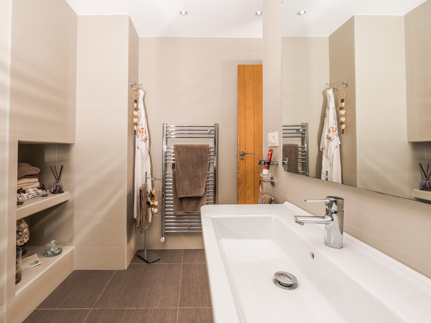 Moderne badkamer met een grote keramische wastafel, ingebouwde wandplanken en een chromen handdoekradiator tegen neutrale beige tegels.