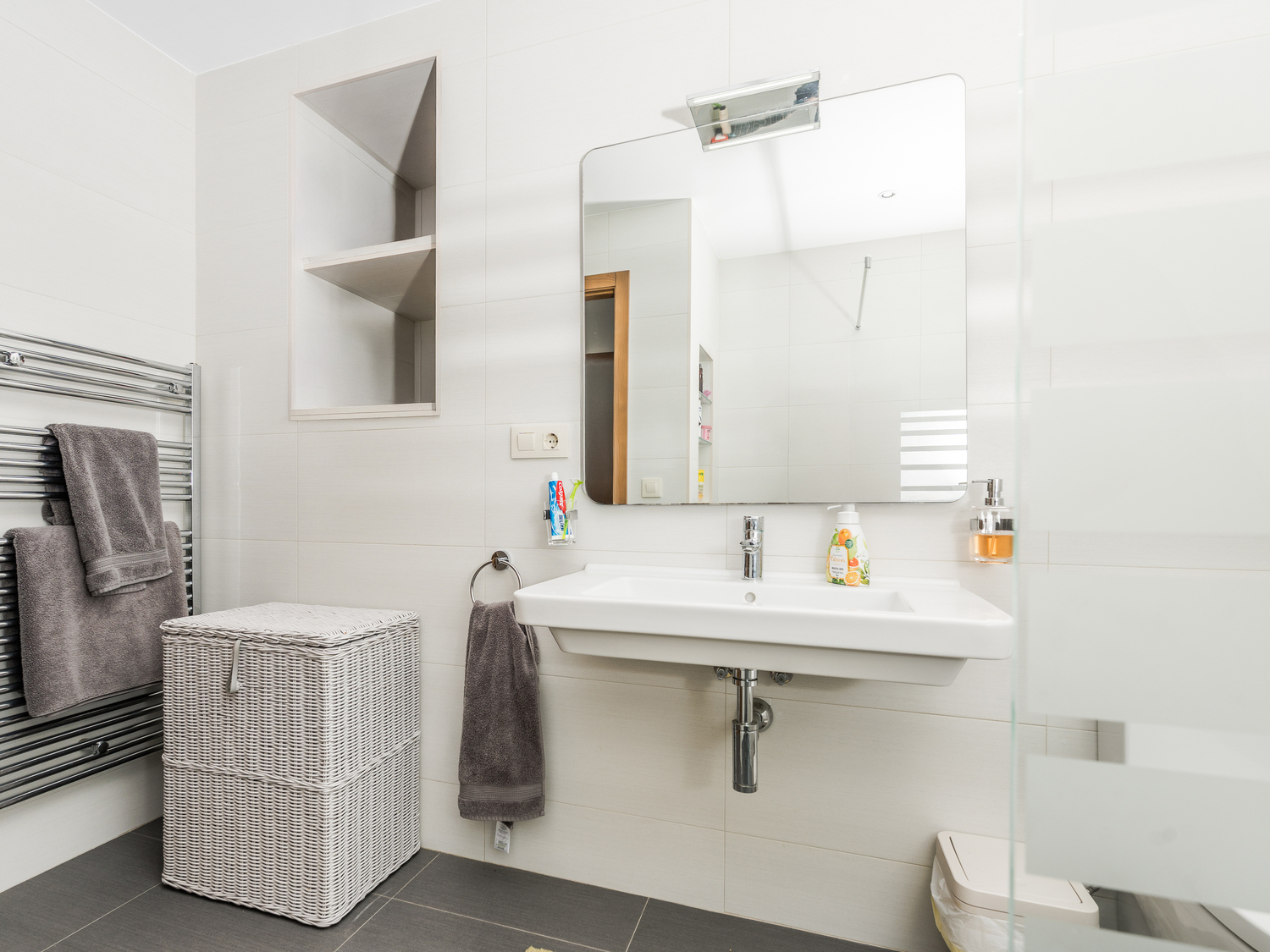 Moderne badkamer met een hangende wastafel, ingebouwde wandnis en chromen handdoekradiator tegen witte wandtegels en grijze vloer.