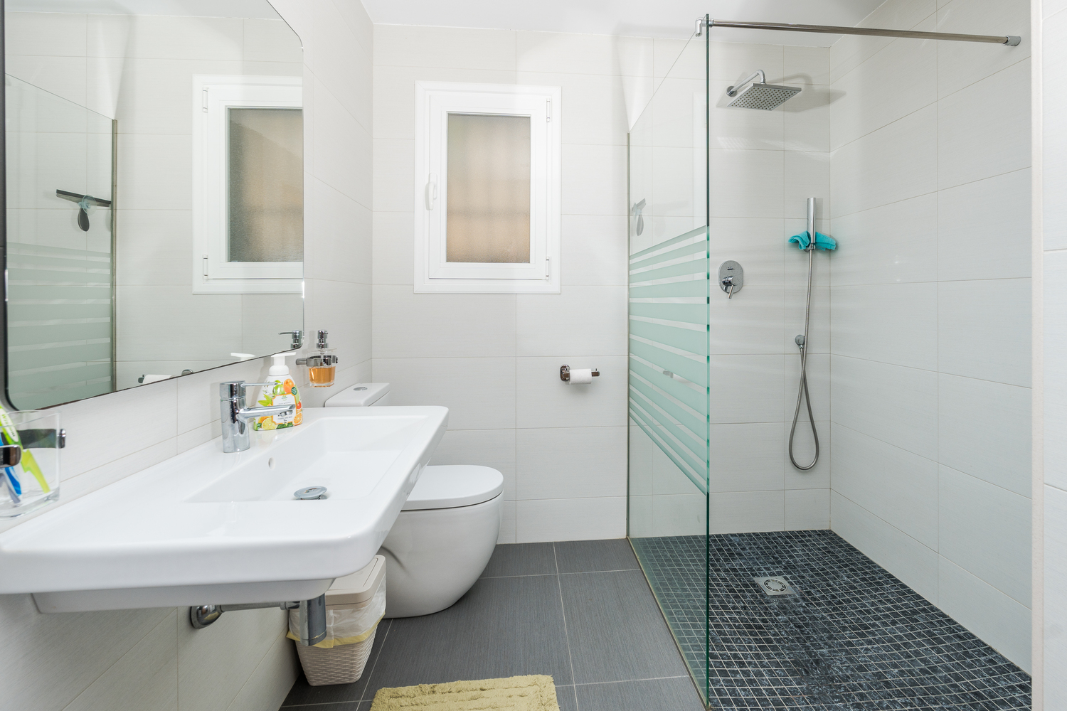 Moderne badkamer met inloopdouche, glazen wand en donkere mozaïekvloer. Voorzien van een zwevend wastafelmeubel en witte wandtegels met natuurlijk lichtinval.