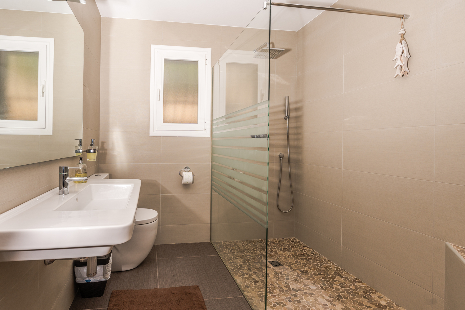 Moderne badkamer met inloopdouche voorzien van kiezelvloer, glazen wand en neutrale betegeling. Inclusief hangend wastafelmeubel en matglas raam.