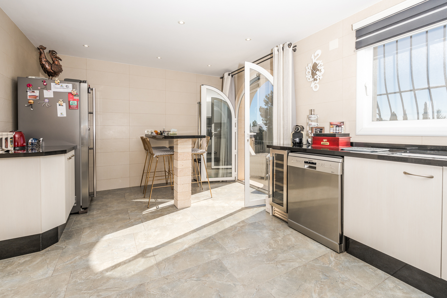 Moderne keuken met witte kasten, zwart granieten werkbladen en een ontbijtbar. Voorzien van rvs-apparatuur en een ronde glazen deur die toegang biedt tot het terras.