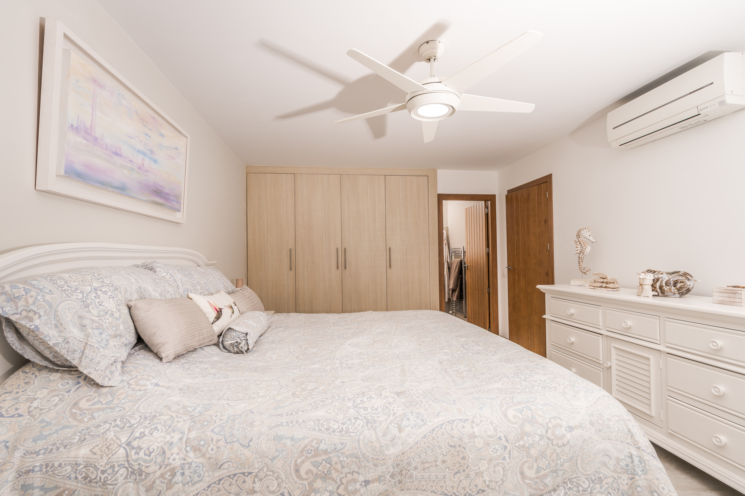 Moderne slaapkamer met ingebouwde licht houten kasten, een witte commode en plafondventilator. Voorzien van wandairconditioning en toegang tot de en-suite kamer.