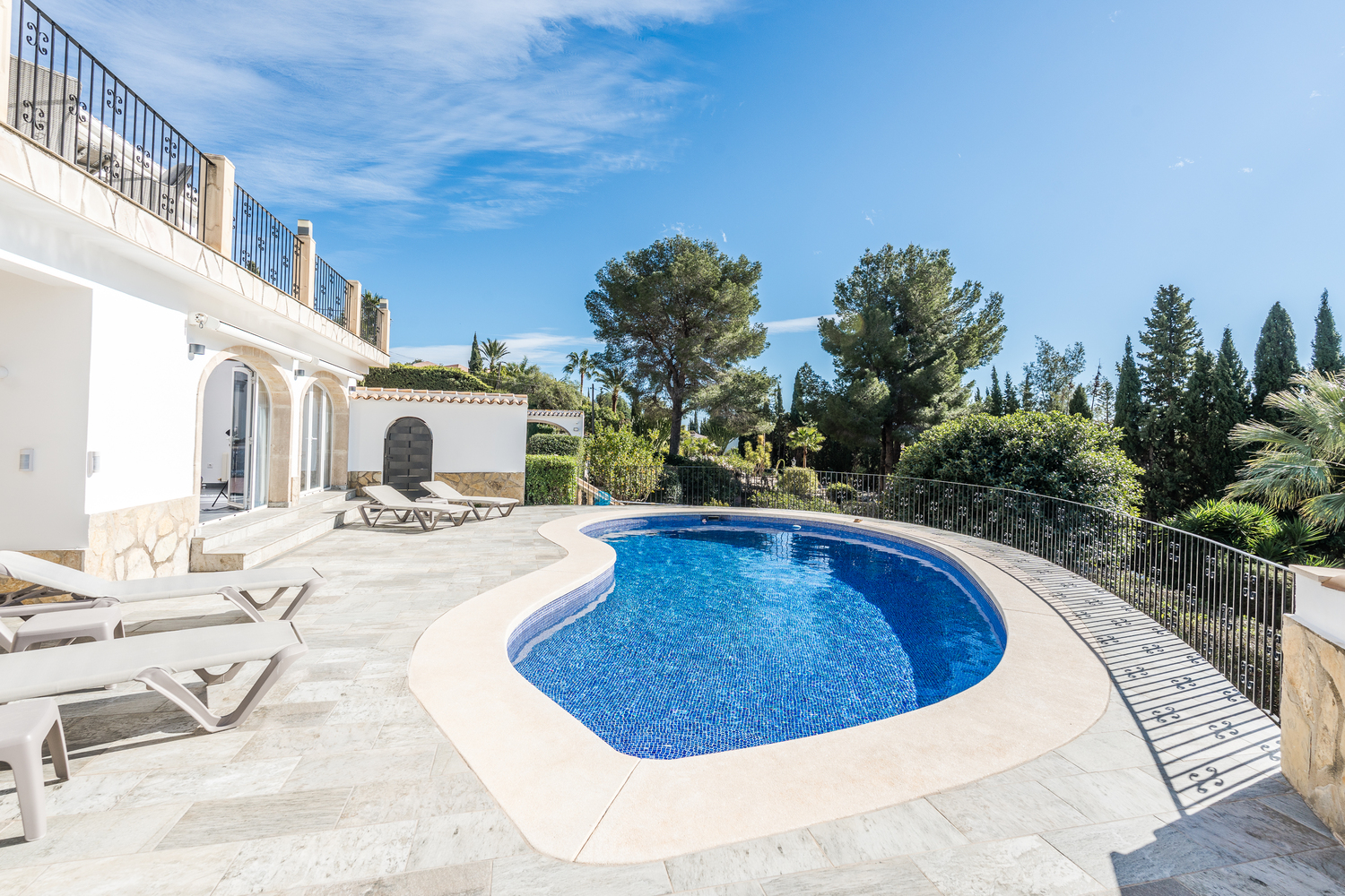 Nier-vormig zwembad omgeven door een licht stenen terras met ligbedden, gelegen tegen een achtergrond van mediterrane bomen en de witte gevel van de villa.