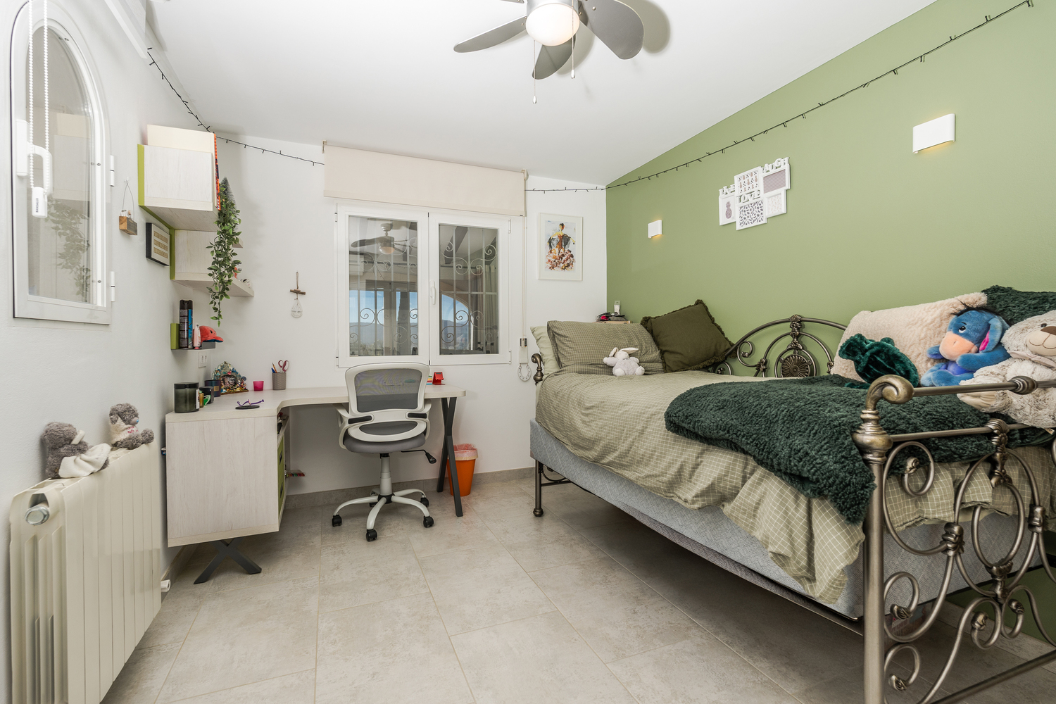 Slaapkamer met een groene accentmuur, een sierlijk metalen bedframe en een geïntegreerde werkplek met bureau en ergonomische stoel. Voorzien van plafondventilator.