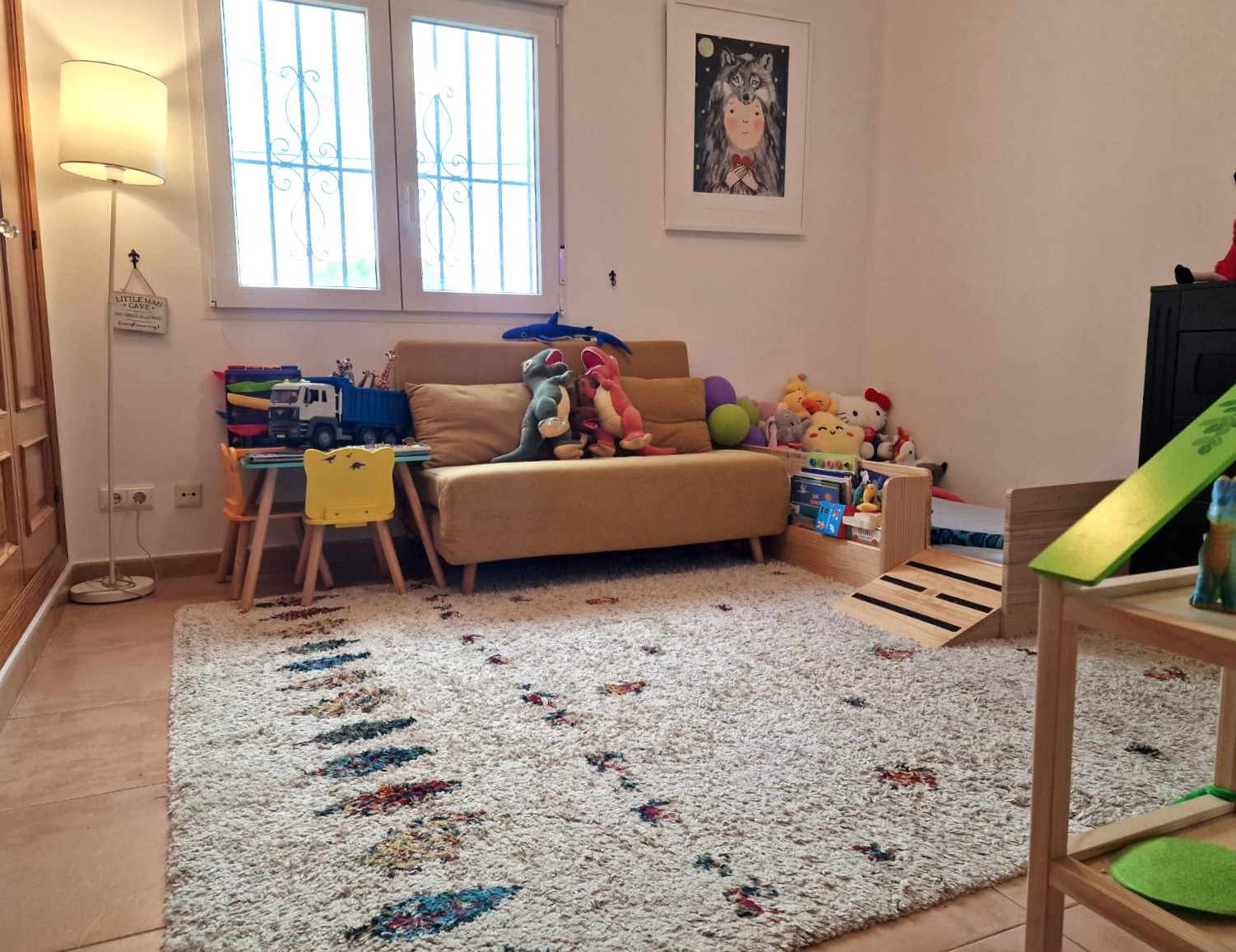 Kinderspeelhoek voorzien van een hoogpolig tapijt, een gele bank en een speeltafel. De ruimte heeft een tegelvloer en lichtinval via een beveiligd venster.