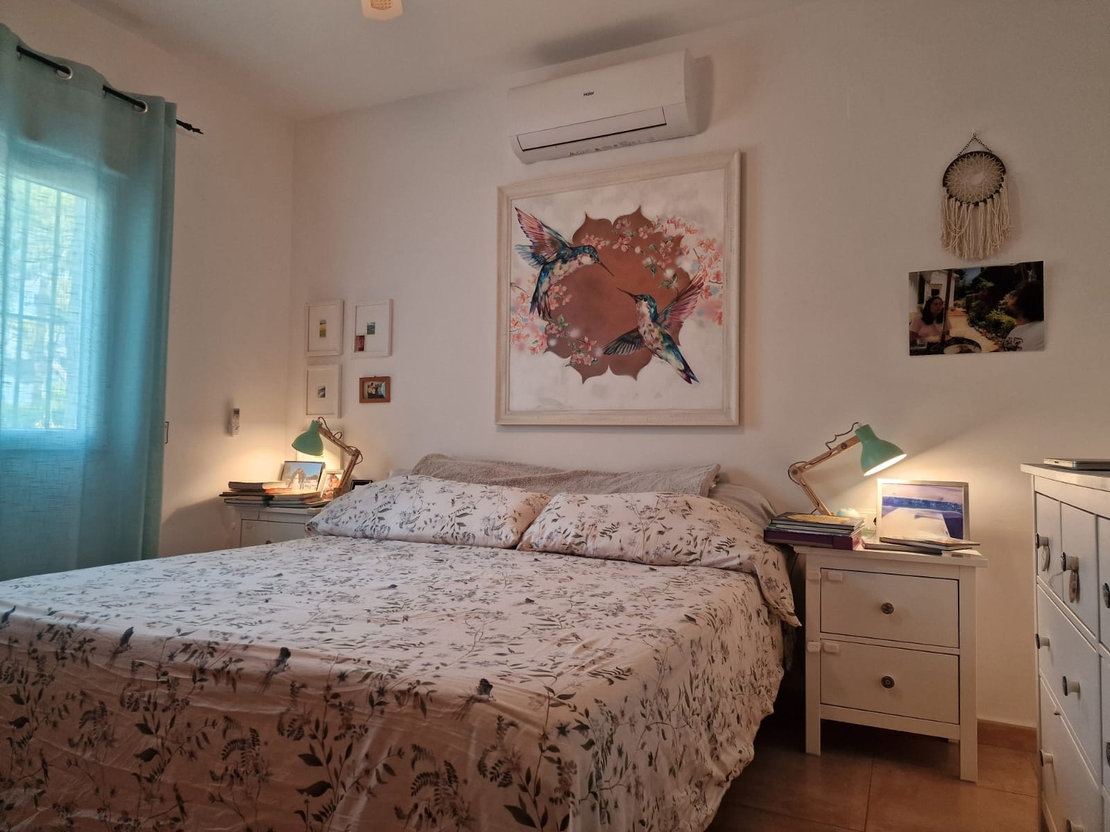 Lichte slaapkamer met tweepersoonsbed, Haier airconditioning, witte nachtkastjes met groene leeslampen en een groot kunstwerk met kolibries boven het bed.