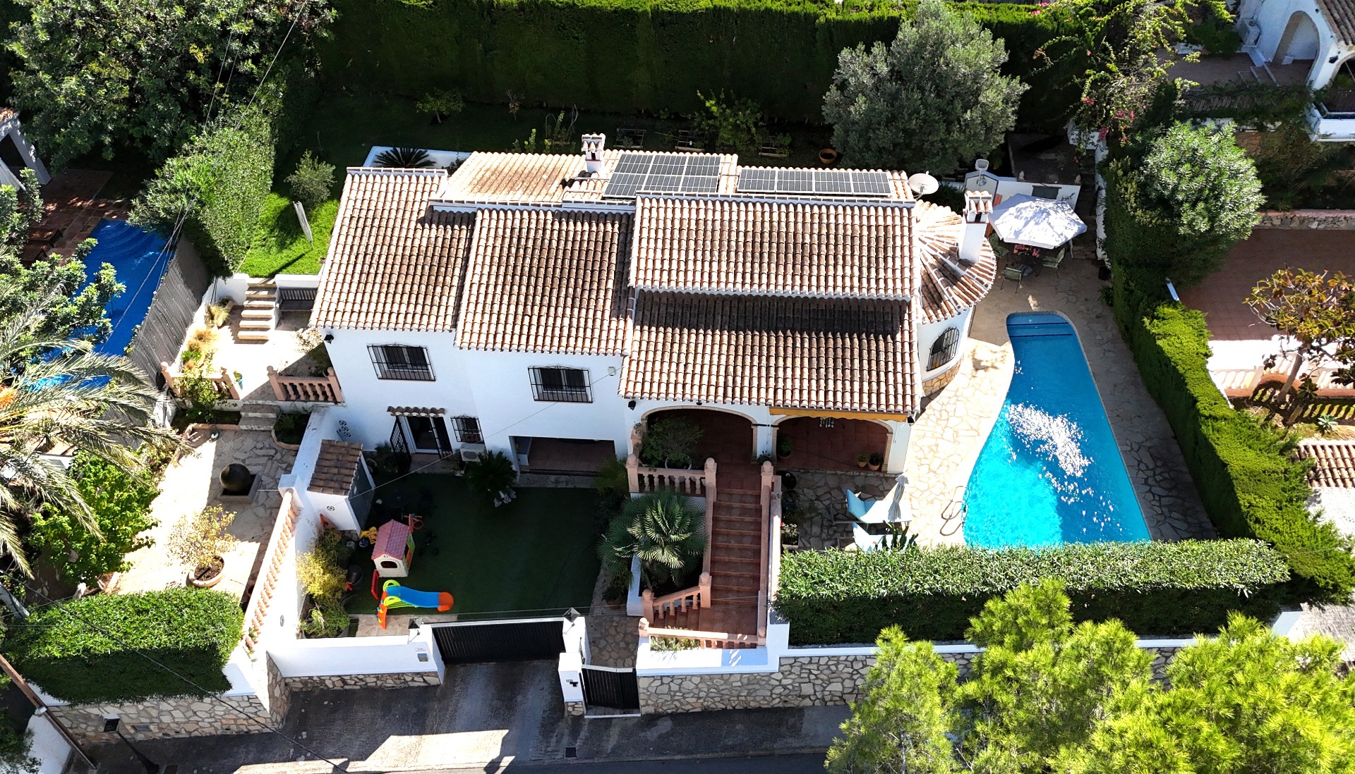 Luchtfoto van deze villa in Toscal met een terracotta dak met zonnepanelen, privézwembad en een kinderspeelplaats in een aangelegde mediterrane tuin.