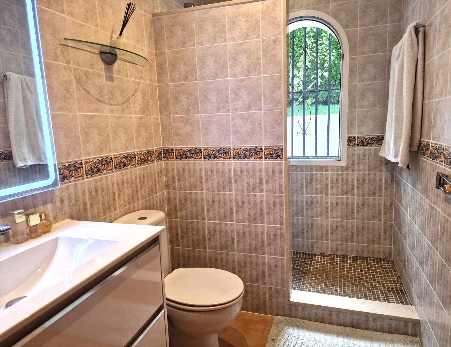 Moderne badkamer met inloopdouche, beige wandtegels met decoratieve rand en een boogvenster dat zorgt voor natuurlijk daglicht.