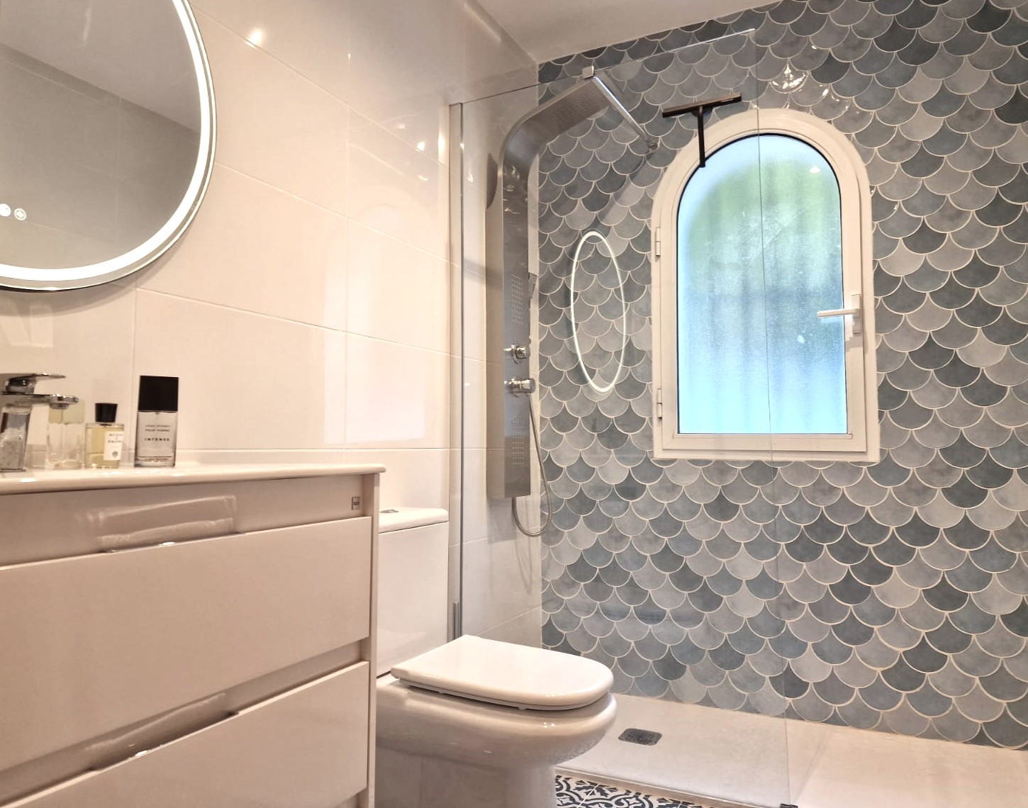 Moderne badkamer met inloopdouche, blauwe schubben tegels, een rondboogvenster met matglas, rvs douchekolom en een verlichte ronde spiegel.