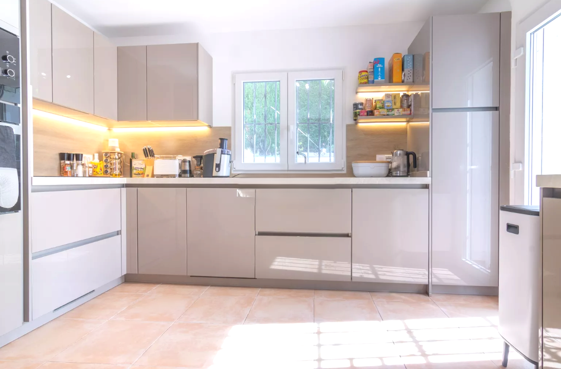 Moderne keuken met hoogglans taupe kasten, witte werkbladen en geïntegreerde LED-verlichting. Voorzien van een terracotta tegelvloer en inbouwapparatuur.