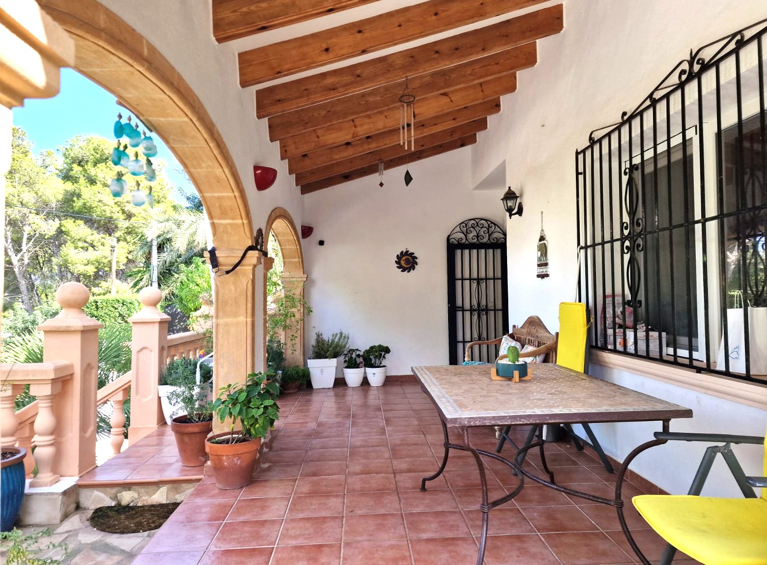 Overdekt terras met Tosca-stenen bogen, houten balkenplafond en terracotta vloer. Buitenruimte met eethoek en uitzicht op de omliggende mediterrane beplanting.