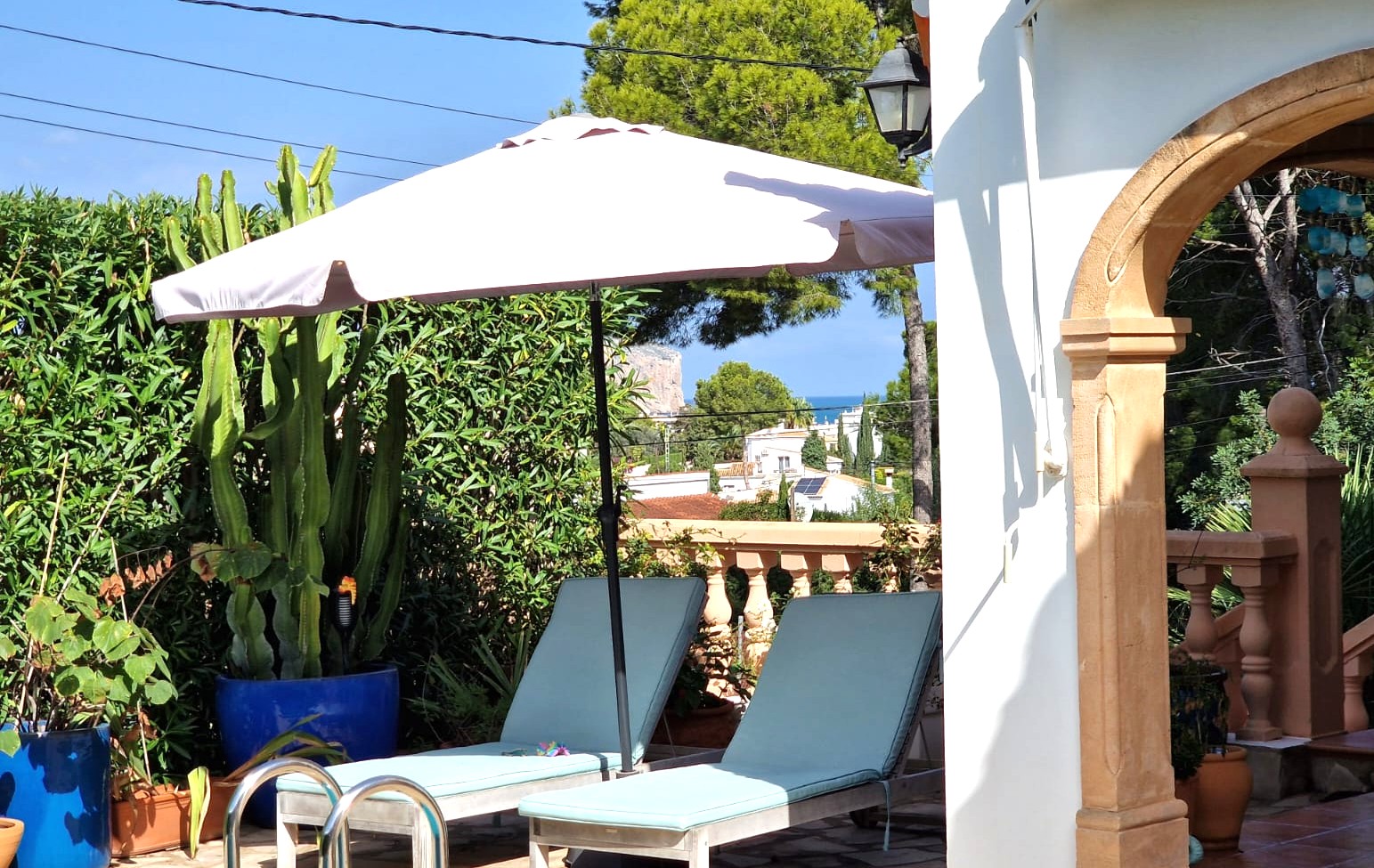 Terras met ligstoelen en parasol bij een stenen boog, omgeven door mediterrane beplanting met uitzicht op zee en Cabo de San Antonio in de verte.