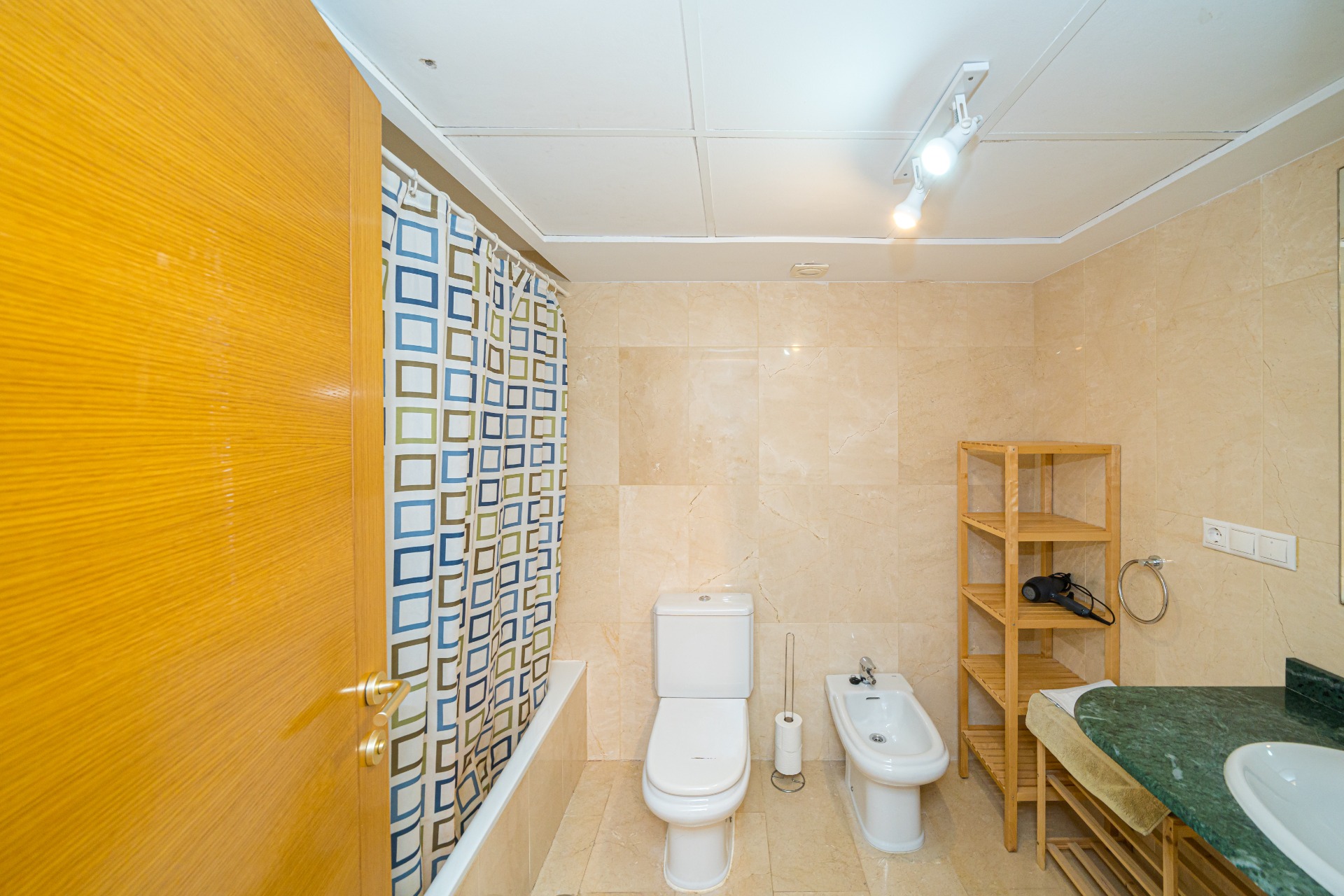 Badkamer met beige marmeren tegels, voorzien van een ligbad, bidet en een groen marmeren wastafelblad met een houten opbergmeubel.