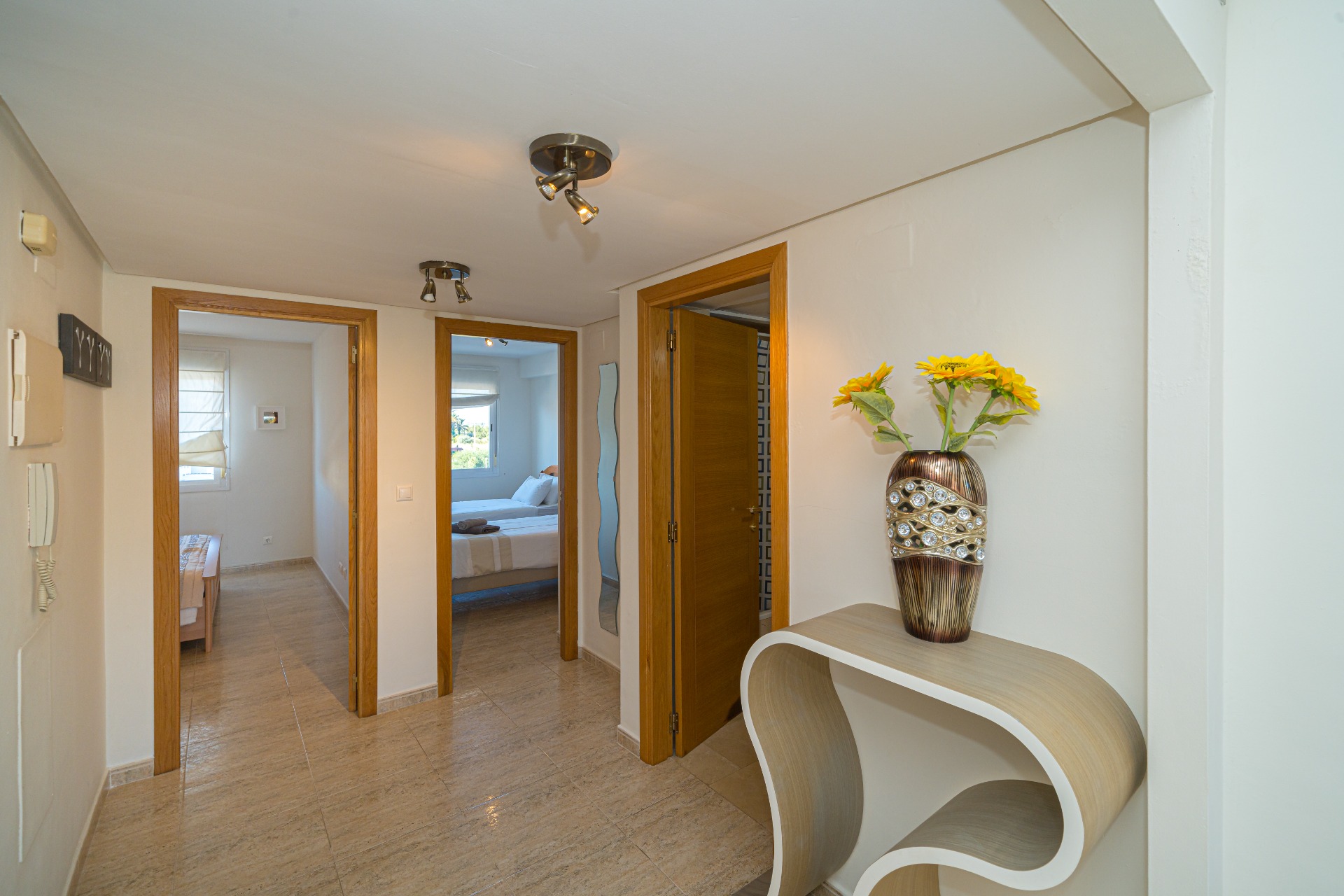 Centrale hal met gepolijste tegelvloer die toegang biedt tot de slaapkamers en badkamer, voorzien van moderne plafondspots en een gebogen consoletafel.