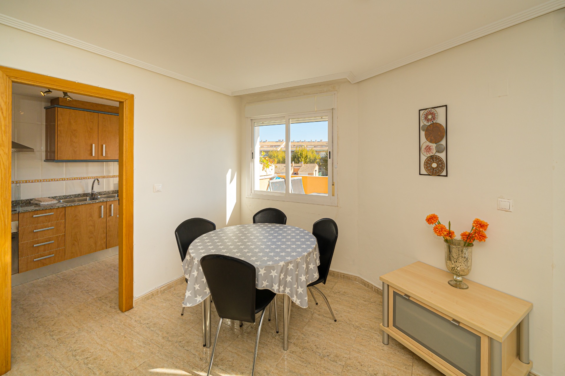 Lichte eethoek met een ronde tafel en tegelvloer, grenzend aan de houten keuken. Het raam biedt natuurlijk licht en uitzicht op de omliggende bebouwing.