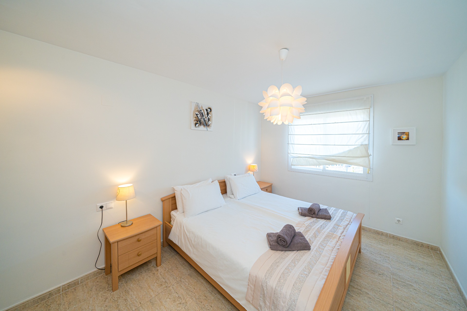 Lichte slaapkamer met licht houten meubilair, een tweepersoonsbed en bijpassende nachtkastjes. Natuurlijk licht valt binnen via het raam met wit rolgordijn op de tegelvloer.