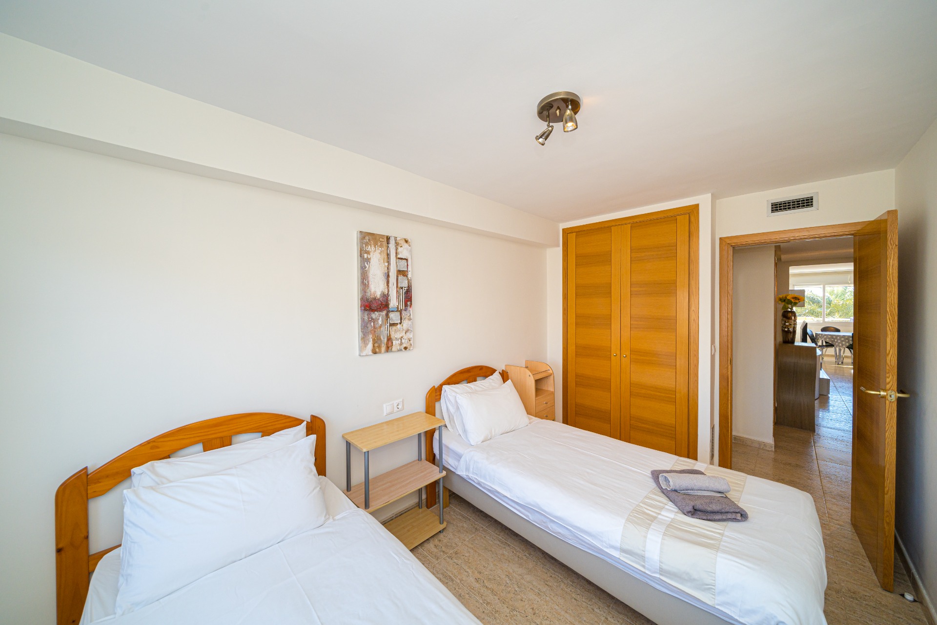 Lichte slaapkamer met twee eenpersoonsbedden, houten hoofdborden, een ingebouwde eikenhouten kast en tegelvloer. Een open deur biedt toegang tot de woonruimte.