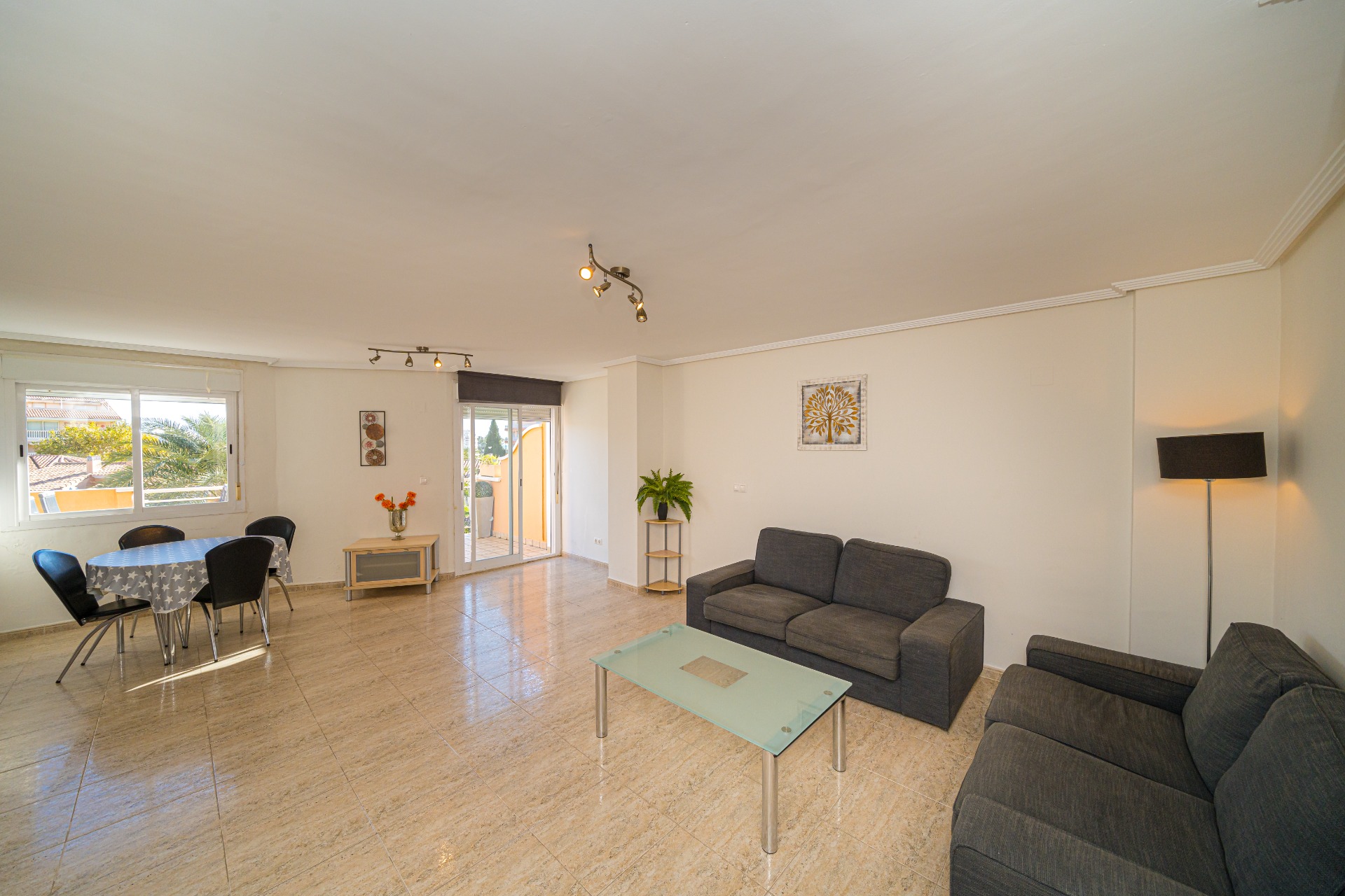 Lichte woon- en eetkamer met een beige tegelvloer, grijze banken en een glazen salontafel. De schuifpui geeft toegang tot het balkon met uitzicht op de omgeving.