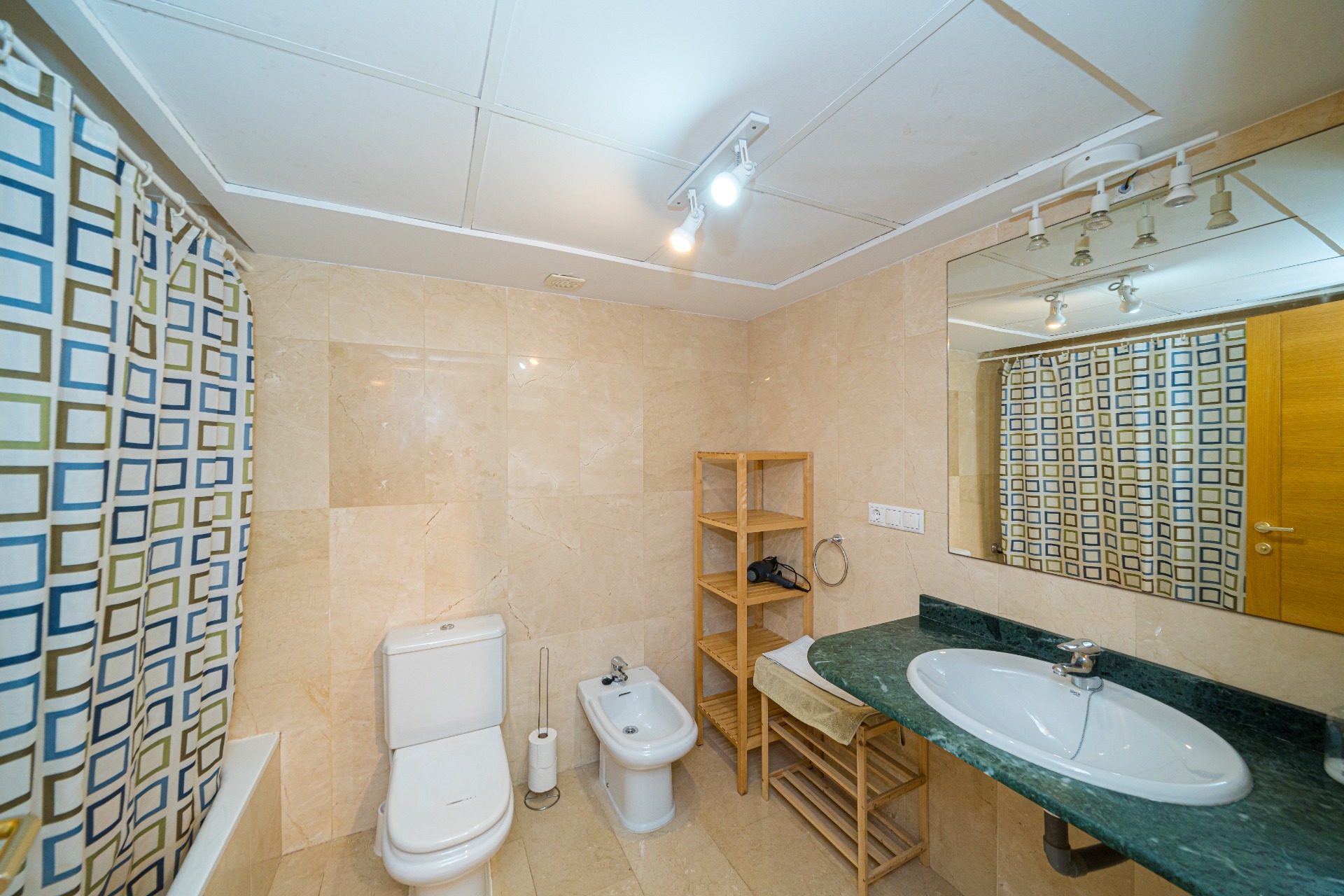 Moderne badkamer met crème marmeren tegels, een groen marmeren wastafelblad en bidet. Voorzien van houten opbergrekken, een ligbad en plafondspots.