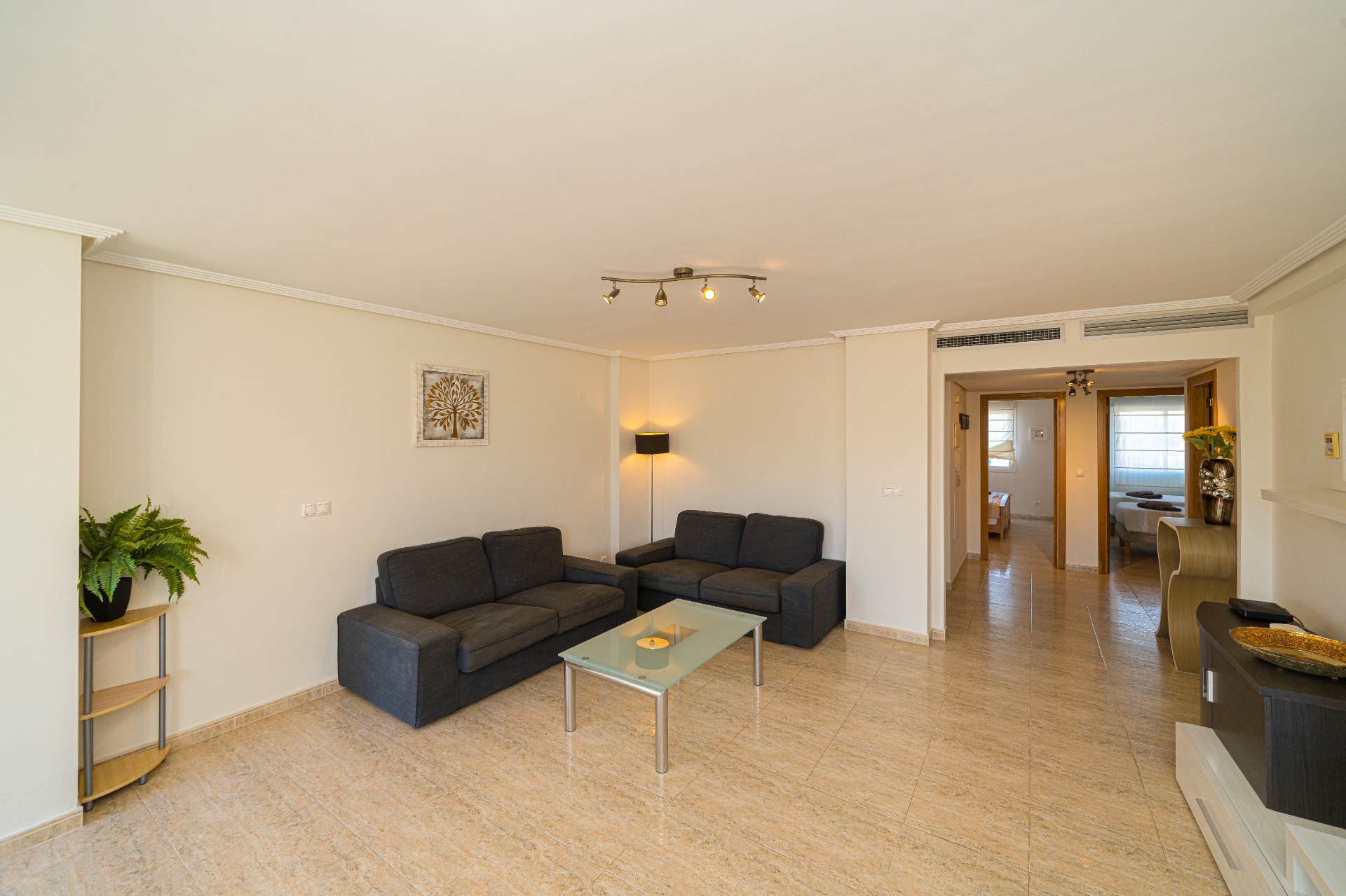Ruime woonkamer met donkergrijze banken, een glazen salontafel en gepolijste tegelvloer. De open indeling biedt toegang tot de gang naar de slaapkamers.