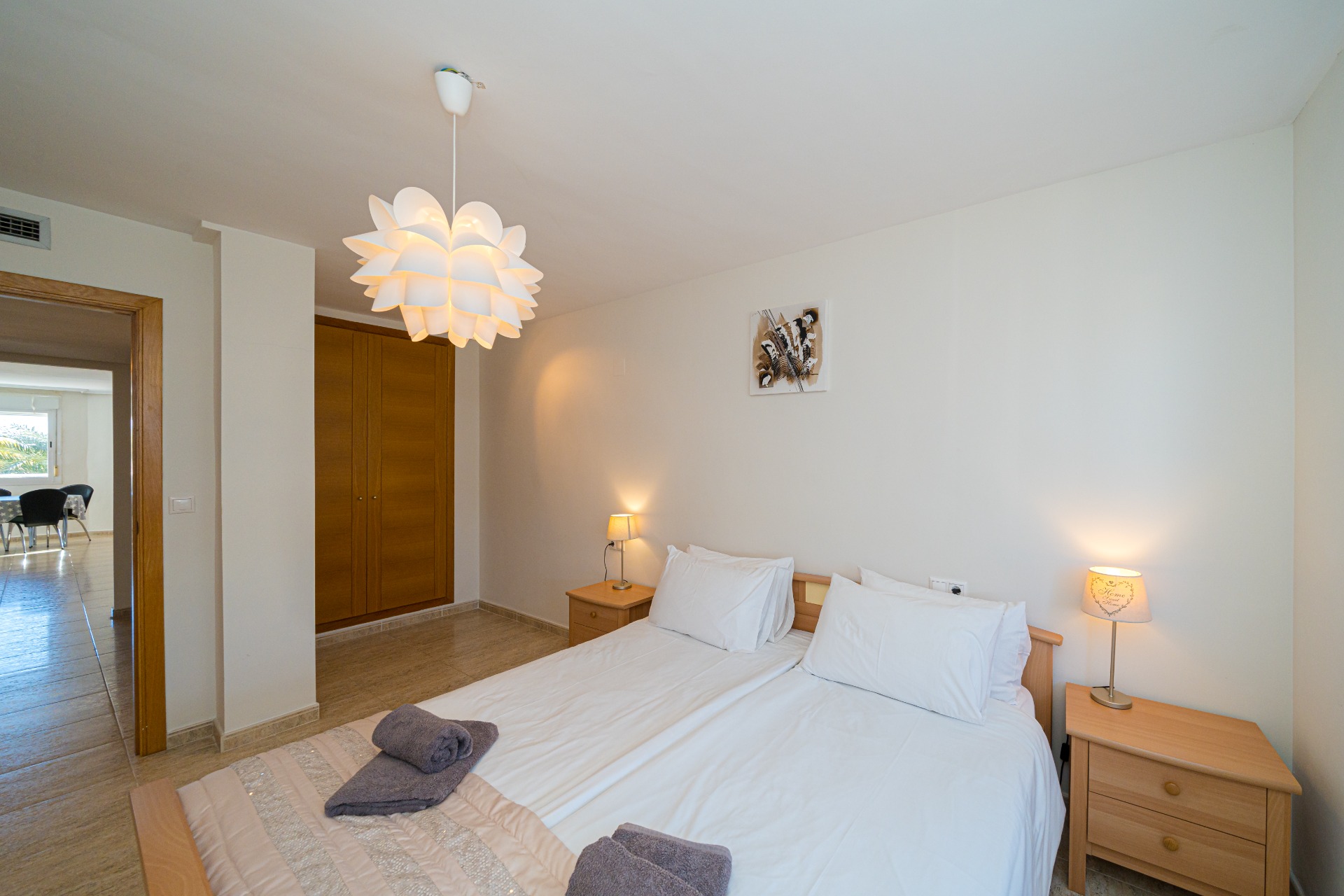 Slaapkamer met twee bedden, houten inbouwkasten en een decoratieve hanglamp. De deuropening biedt een doorkijk naar de aangrenzende lichte eetkamer.