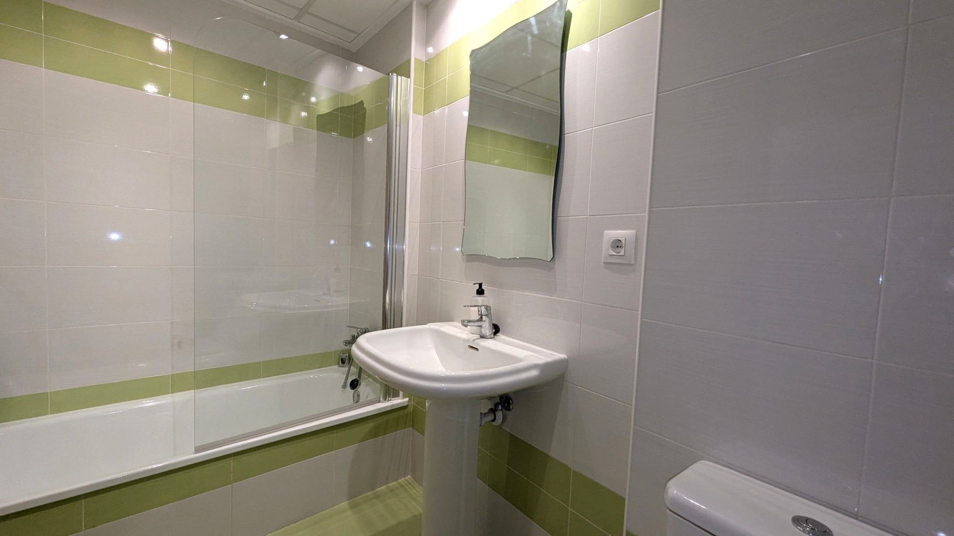 Badkamer met ligbad en glazen douchescherm, kolomwastafel en wandtegels in wit en limoengroen met een golvende spiegel.
