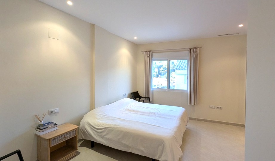 Lichte slaapkamer in neutrale tinten met inbouwspots in het plafond en een raam voorzien van rolluiken. Voorzien van een tegelvloer en een houten nachtkastje.