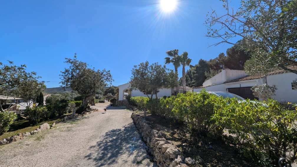Brede oprit van grind geflankeerd door traditionele stenen muren die leidt naar het hoofdhuis in finca-stijl, omgeven door mediterrane beplanting en palmbomen.