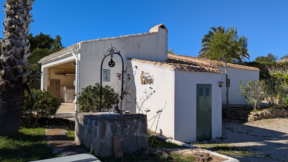 Buitenaanzicht van een traditioneel mediterraan bijgebouw met witte gevels, pannendak en een decoratieve stenen put met smeedijzeren details.