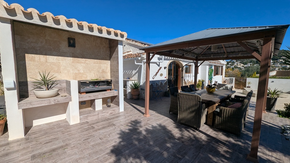 Buitenruimte met een ingebouwde stenen barbecue en een ruim terras onder een pergola, ingericht met rijk gevlochten stoelen op een houtlook tegelvloer.
