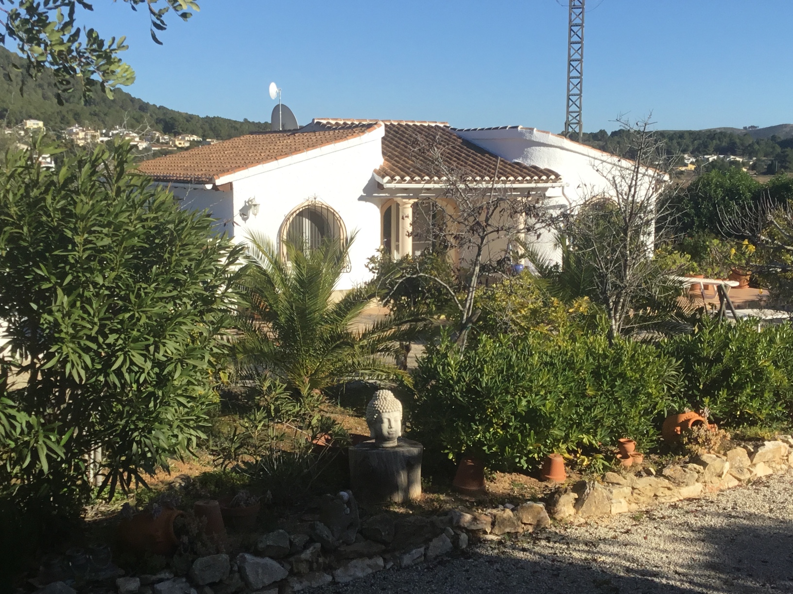 Exterieur van de witgekalkte mediterrane villa met boogvensters, terracotta dakpannen en een aangelegde tuin met palmbomen en een Boeddhabeeld.