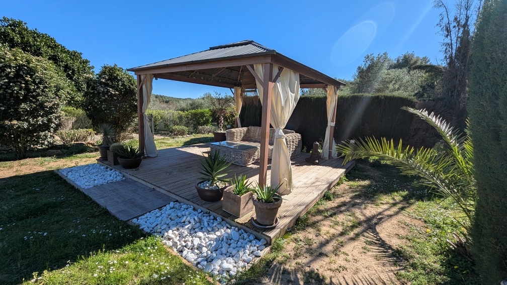 Houten tuinpaviljoen op een vlonderterras met wicker loungeset, crèmekleurige gordijnen en witte sierkeien, gelegen in een rustige mediterrane tuin in Jávea.