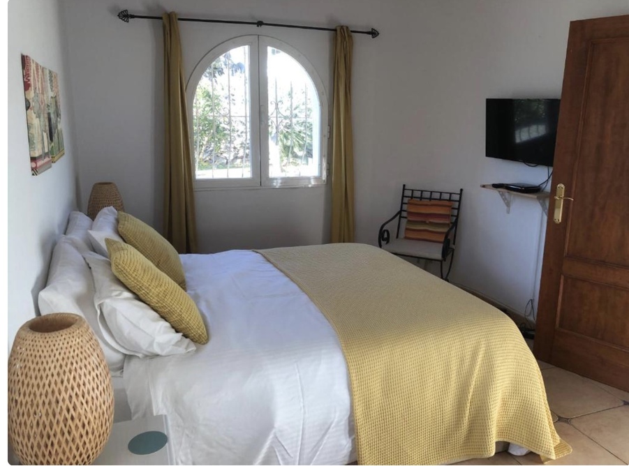 Lichtrijke slaapkamer met een boogvenster, terracotta vloertegels en een wand-tv. De kamer is ingericht met mosterdgele accenten en gevlochten nachtlampen.
