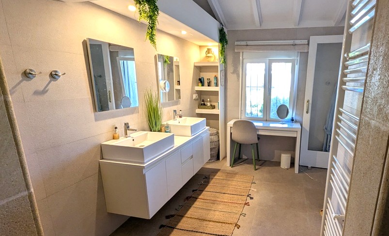 Moderne badkamer met dubbele wastafel, zwevend meubel en ingebouwde kasten. Inclusief een kaptafel bij het raam en een plafond met witte houten balken.