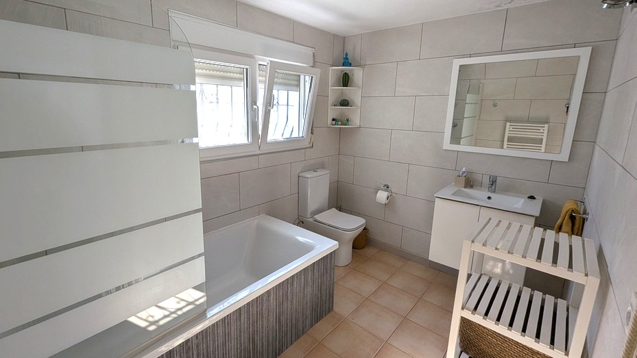 Moderne badkamer met ingebouwd ligbad, matglazen douchescherm, wit wastafelmeubel en grootformaat wandtegels in neutrale kleurstelling.