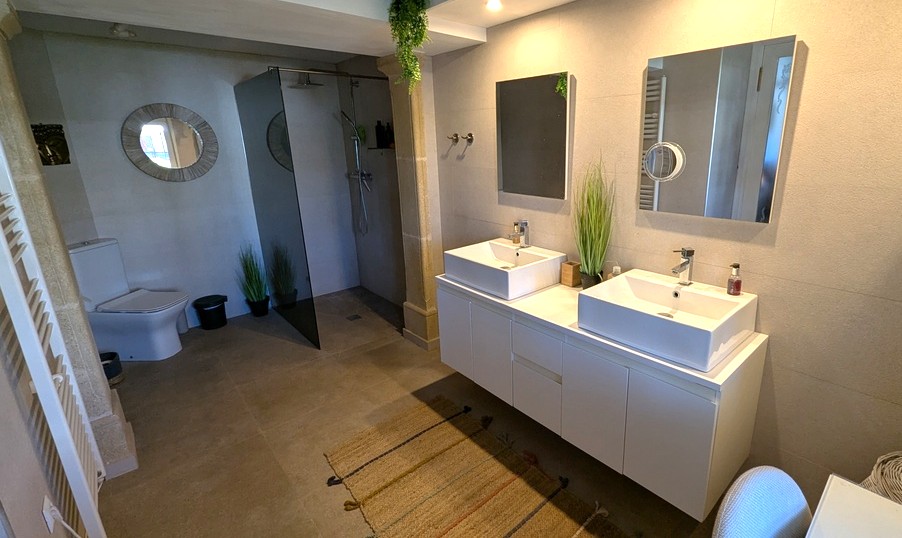 Moderne badkamer voorzien van een dubbele wastafel, inloopdouche met getinte glaswand en neutrale tegelafwerking met authentieke stenen details.
