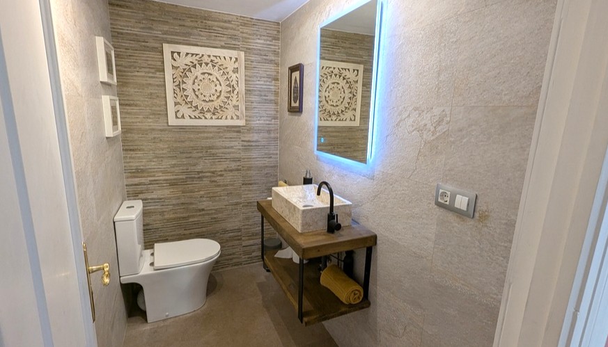 Moderne badkamer voorzien van een natuurstenen waskom op een zwevend houten meubel, een verlichte LED-spiegel en wandtegels met een steenstructuur.