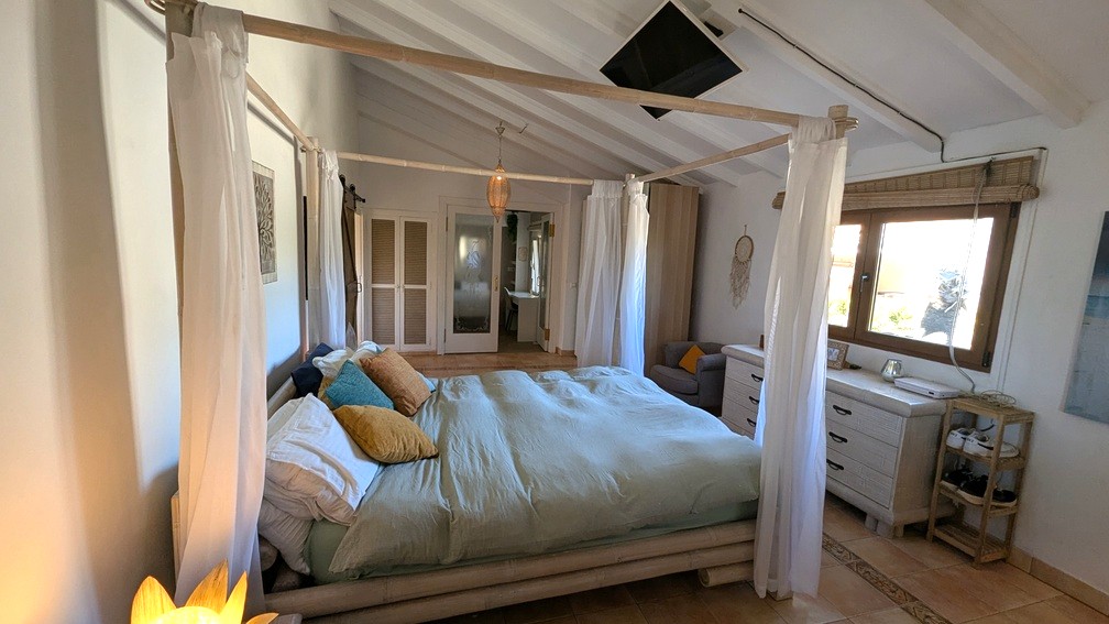 Slaapkamer met bamboe hemelbed, wit geschilderd balkenplafond en terracotta vloertegels. Inclusief aan het plafond gemonteerde tv en inbouwkasten.